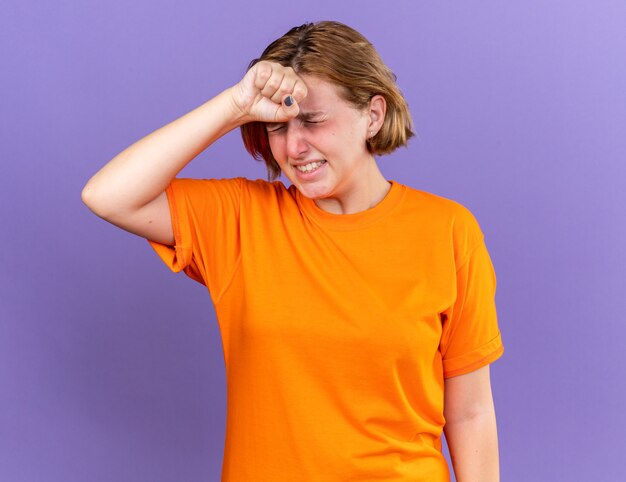 Mulher jovem insalubre com uma camiseta laranja se sentindo péssima ao tocar na testa enquanto se sente tonta com gripe