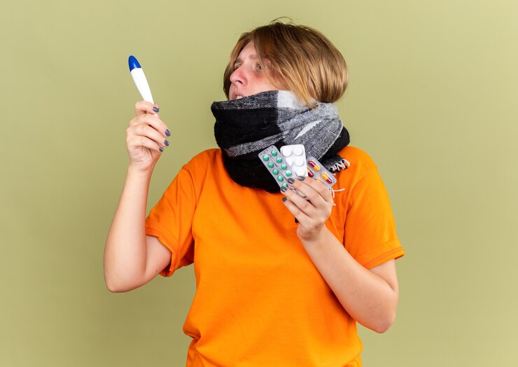 Foto grátis mulher jovem insalubre em uma camiseta laranja com um lenço quente no pescoço sentindo uma terrível dor de gripe segurando comprimidos para verificar a temperatura usando um termômetro e parece preocupada na parede verde