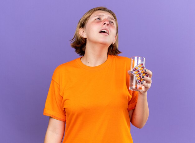 Mulher jovem insalubre em uma camiseta laranja se sentindo mal segurando um copo de água e comprimidos contra a gripe espirrando