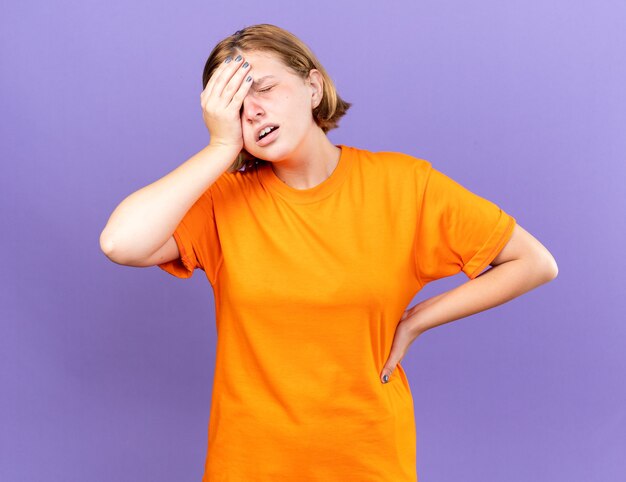 Mulher jovem insalubre em uma camiseta laranja se sentindo mal tocando sua testa enquanto se sente tonta com gripe em pé sobre uma parede roxa