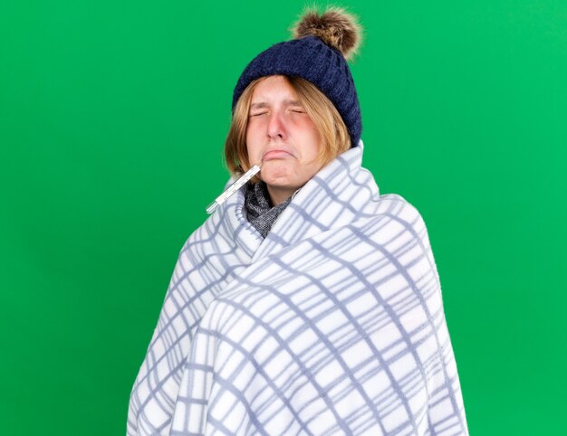 Mulher jovem insalubre enrolada em um cobertor, usando um chapéu, medindo a temperatura do corpo usando um termômetro, sentindo-se doente, sofrendo de gripe, tendo febre em pé sobre uma parede verde