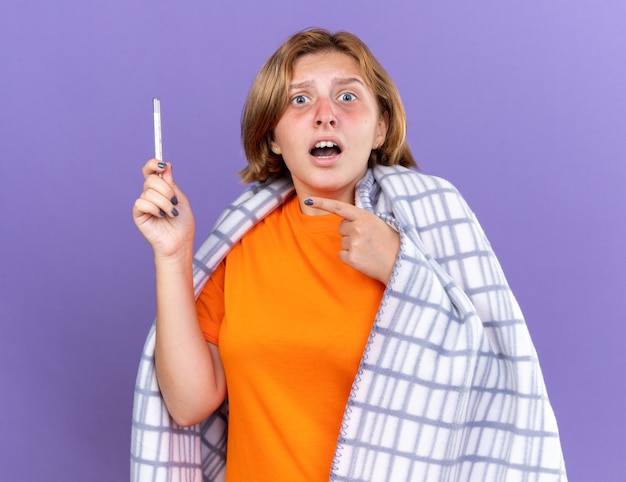 Mulher jovem insalubre envolta em um cobertor quente sentindo-se doente, sofrendo de gripe, tendo febre medindo a temperatura usando um termômetro apontando para ele, parecendo preocupada em pé sobre a parede roxa
