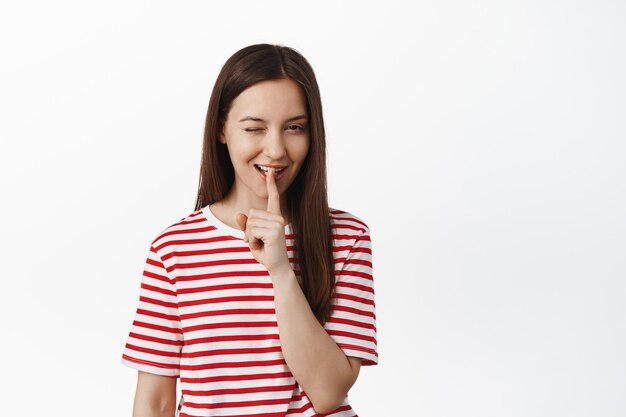 Mulher jovem insolente pisca e silencia, faz um gesto shh com o dedo perto dos lábios enquanto sorri, tem surpresa, insinuando algo, de pé em camiseta vermelha contra fundo branco