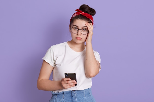 Mulher jovem isolada sobre uma parede roxa com expressão facial preocupada, segurando um telefone inteligente quebrado, vestindo uma camiseta casual branca e faixa de cabelo vermelha