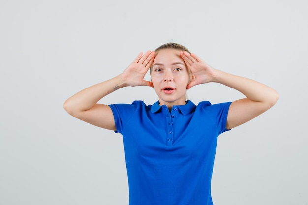 Mulher jovem levantando as mãos para ver claramente em uma camiseta azul e parecendo curiosa
