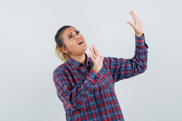 Mulher jovem, levantando as mãos, tentando impedir algo em uma camisa xadrez e olhando com medo, vista frontal.