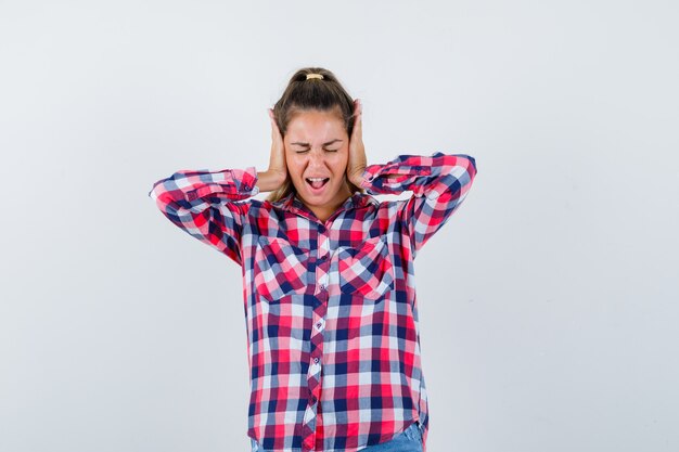 Mulher jovem, mantendo as mãos nas orelhas na camisa casual e parecendo irritada, vista frontal.