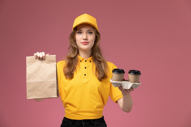 Mulher jovem mensageira de uniforme amarelo segurando copos de plástico de café marrom e um pacote de comida na mesa rosa-escuro. Serviço de entrega de uniforme