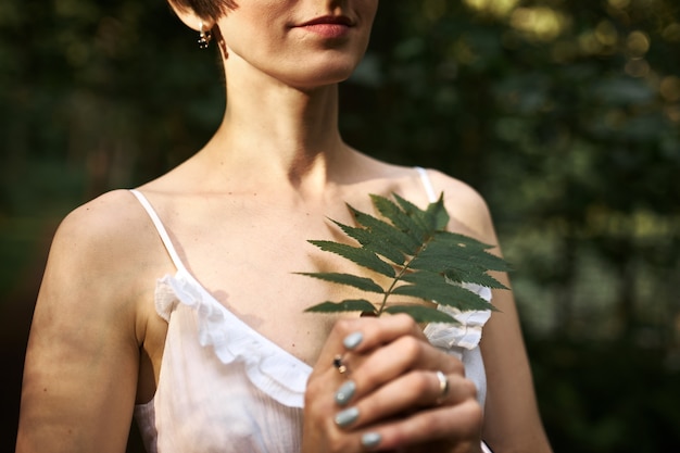 Mulher jovem misteriosa irreconhecível com penteado curto e pele pálida, caminhando na floresta sozinha, segurando uma folha de samambaia verde.