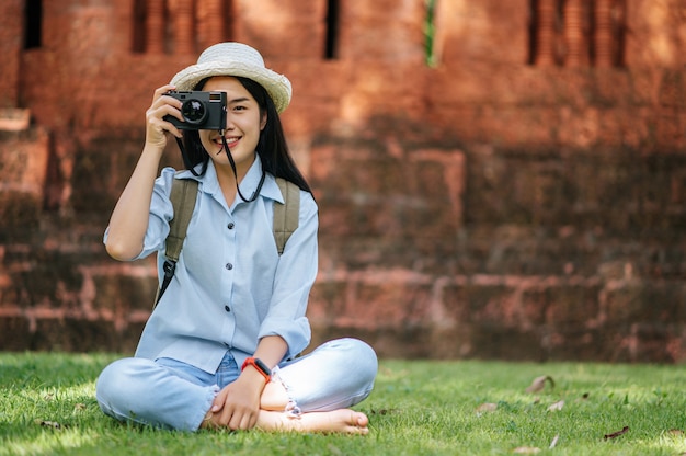 Mulher jovem mochileira asiática usando chapéu sorrindo enquanto viaja em um local histórico, ela sentada na grama para relaxar e usar a câmera para tirar uma foto com felicidade