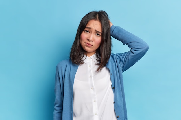 Mulher jovem morena mal-humorada decepcionada com aparência oriental coça a cabeça carranca rosto parece infeliz para a câmera usa camisa branca e jumper azul posa interior. conceito de emoções negativas