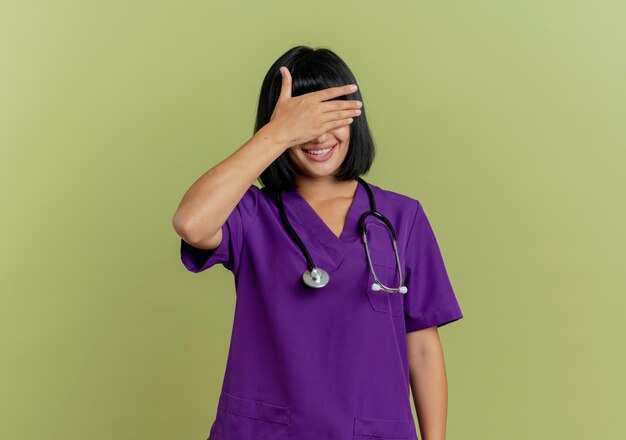 Mulher jovem morena sorridente de uniforme com estetoscópio fecha os olhos com a mão isolada em fundo verde oliva com espaço de cópia