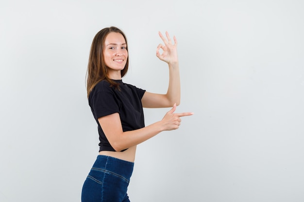 Mulher jovem mostrando ok e disparando gesto de arma em blusa preta, calça e parecendo feliz