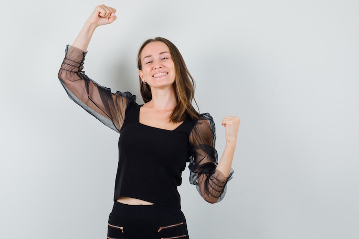Foto grátis mulher jovem mostrando os músculos e sorrindo com uma blusa preta e calça preta e parecendo confiante