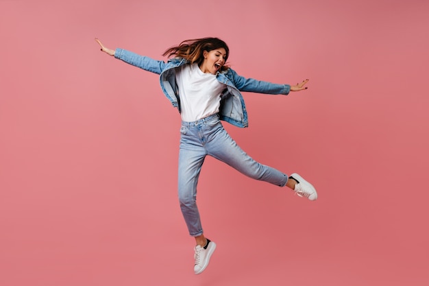 Mulher jovem na moda pulando no fundo rosa. Visão de comprimento total da despreocupada modelo feminino em roupa de jeans.