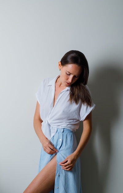 Mulher jovem olhando para baixo e posando para a frente com uma camiseta branca e saia azul claro e parecendo charmosa