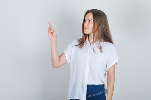 Mulher jovem olhando para longe com o dedo para cima em uma camiseta branca, jeans e parecendo feliz