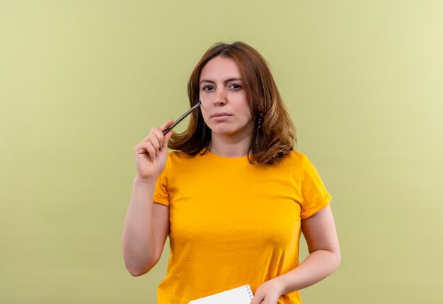 Mulher jovem pensativa e casual segurando uma caneta e um bloco de notas na parede verde isolada com espaço de cópia