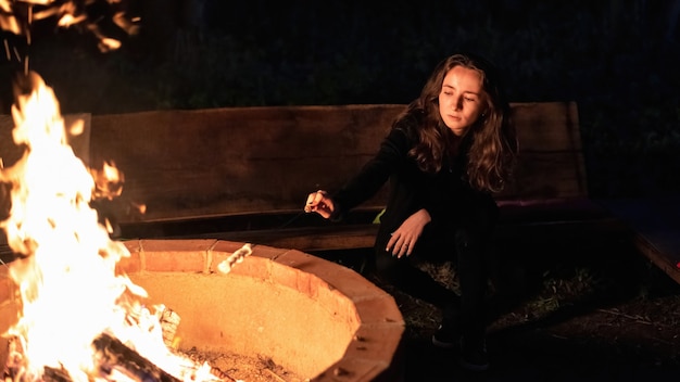 Mulher jovem perto de uma fogueira em glamping, à noite. fritando marshmallow