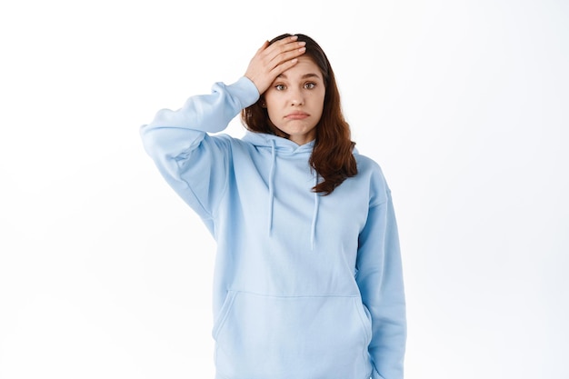 Mulher jovem perturbada se esqueceu ou se lembra de algo, segurando a mão na testa, fazendo facepalm e fazendo beicinho incomodada, tendo problemas, em pé contra um fundo branco