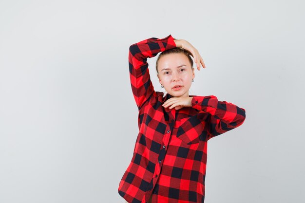 Mulher jovem posando com as mãos na cabeça e embaixo do queixo, com uma camisa xadrez e aparência elegante