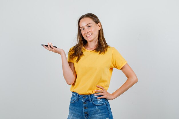 Mulher jovem posando enquanto segura o smartphone em uma camiseta, shorts e parece feliz