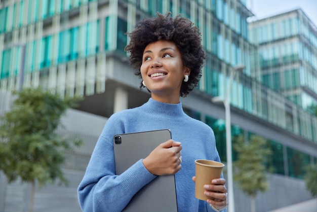 Foto grátis mulher jovem positiva segura tablet digital usa moderno gadget bebidas café para viagem sorrisos feliz usa poses casuais de jumper azul em ambiente urbano retorna de estudar. conceito de pessoas e estilo de vida