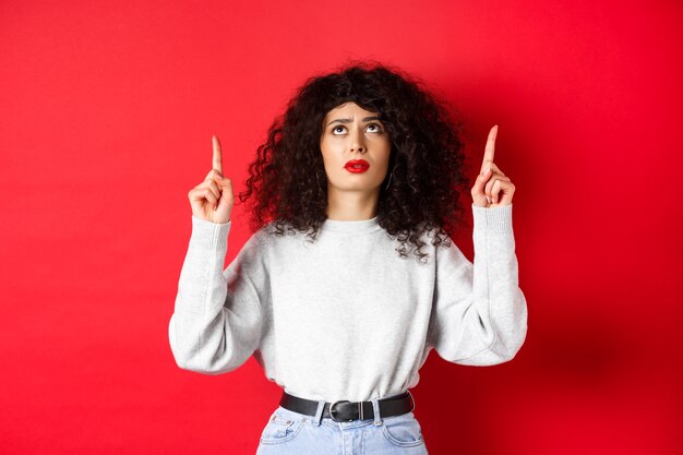 Mulher jovem preocupada com cabelo encaracolado, carrancuda e parecendo em dúvida, apontando para cima com uma cara hesitante ou preocupada, de pé sobre fundo vermelho.