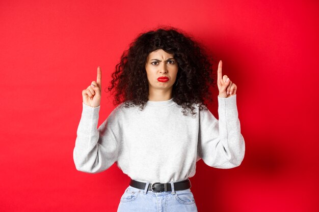 Mulher jovem rabugenta com cabelos cacheados franzindo a testa e fazendo careta insatisfeita, apontando o dedo para algo ruim, reclamando da empresa, fundo vermelho