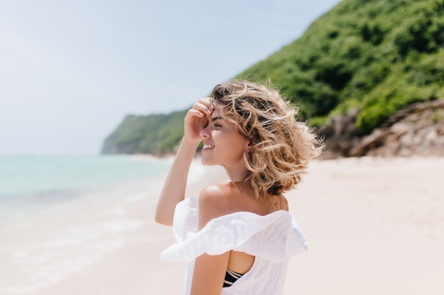 Mulher jovem refinada com cabelo curto e claro, olhando para o mar. Retrato ao ar livre de uma linda mulher bronzeada, caminhando na praia.