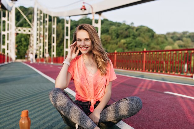 Mulher jovem romântica em camiseta rosa posando antes do treino. Foto ao ar livre de modelo feminino inspirado sentado com as pernas cruzadas na pista de concreto.