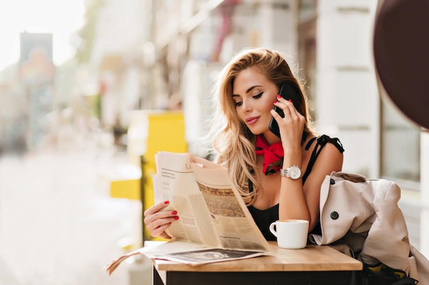 Mulher jovem satisfeita com a pele bronzeada segurando o smartphone e lendo o artigo durante o intervalo para o café. Retrato ao ar livre da menina sorridente no relógio de pulso, falando no telefone, no café, pela manhã.
