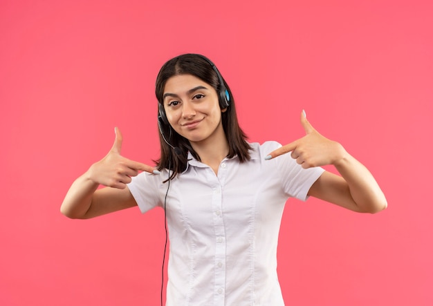 Mulher jovem satisfeita com uma camisa branca e fones de ouvido com um microfone apontando os dedos para si mesma em pé sobre a parede rosa