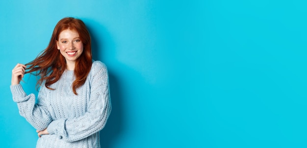 Foto grátis mulher jovem sedutora com cabelo ruivo brincando com o cabelo e sorrindo de pé no suéter contra as costas azuis