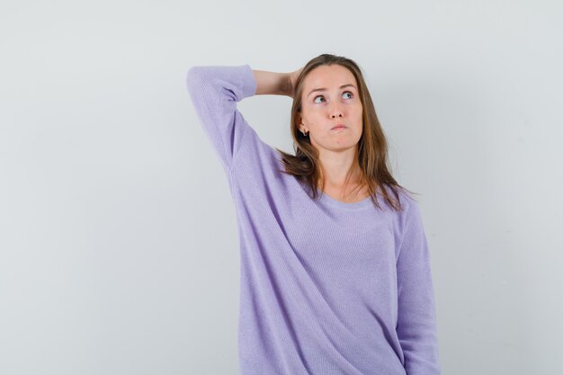 Mulher jovem segurando a mão atrás da cabeça enquanto olha para o lado com uma blusa lilás e parece pensativa