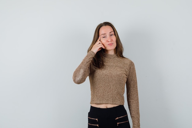 Mulher jovem segurando a mão na bochecha e posando na frente com um suéter dourado dourado e calça preta e está linda