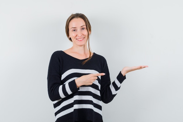 Mulher jovem segurando algo imaginário e apontando para ele, em malha listrada e calça preta, parecendo feliz