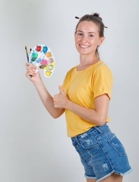 Mulher jovem segurando ferramentas de pintura com o polegar para cima em uma camiseta amarela, shorts jeans e parecendo alegre
