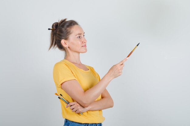 Mulher jovem segurando ferramentas de pintura e olhando para longe em uma camiseta amarela