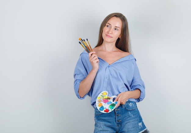 Mulher jovem segurando ferramentas de pintura em uma camiseta, shorts e parecendo feliz
