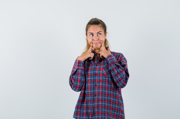 Mulher jovem segurando o dedo indicador perto da boca, forçando um sorriso em uma camisa quadriculada e parecendo feliz