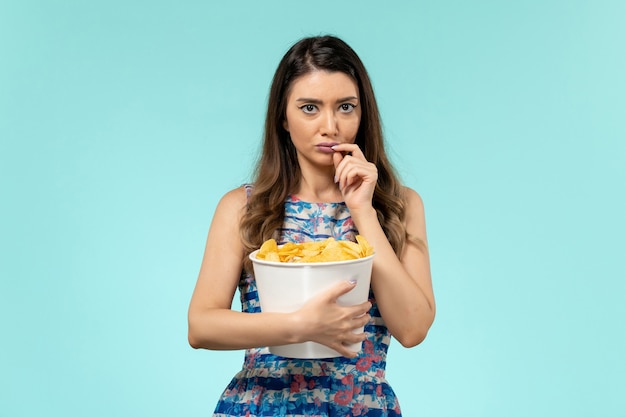 Mulher jovem segurando o pacote com batatas fritas de frente e assistindo filme na superfície azul