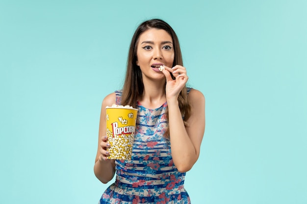 Mulher jovem segurando um pacote de pipoca e comendo na superfície azul de frente