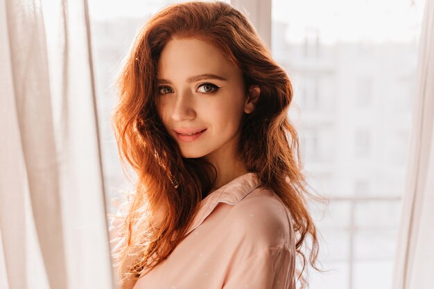 Mulher jovem sensual, com cabelo ruivo em pé perto da janela. Retrato interno de atraente garota branca.