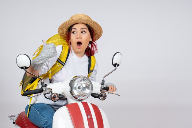 Mulher jovem sentada de frente em uma motocicleta com uma mochila e um chapéu na parede branca