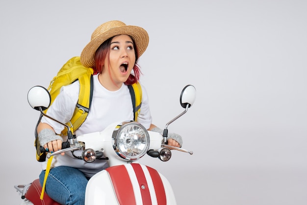 Mulher jovem sentada de frente em uma motocicleta com uma mochila e um chapéu na parede branca