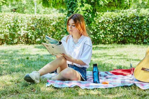 Mulher jovem sentada na grama enquanto lê o diário ao lado do violão