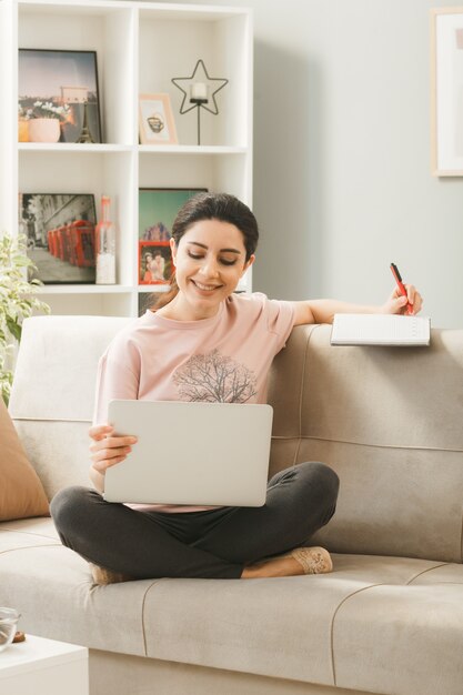 Mulher jovem sentada no sofá, atrás da mesa de centro, segurando e usando um laptop escrevendo em um livro na sala de estar