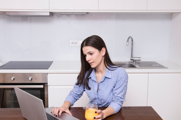 Mulher jovem, sentando, cozinha, segurando, vidro suco, usando computador portátil