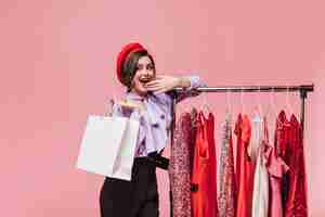 Foto grátis mulher jovem sorri e cobre a boca enquanto faz compras. senhora de boina posa perto de carrinho com vestidos elegantes.